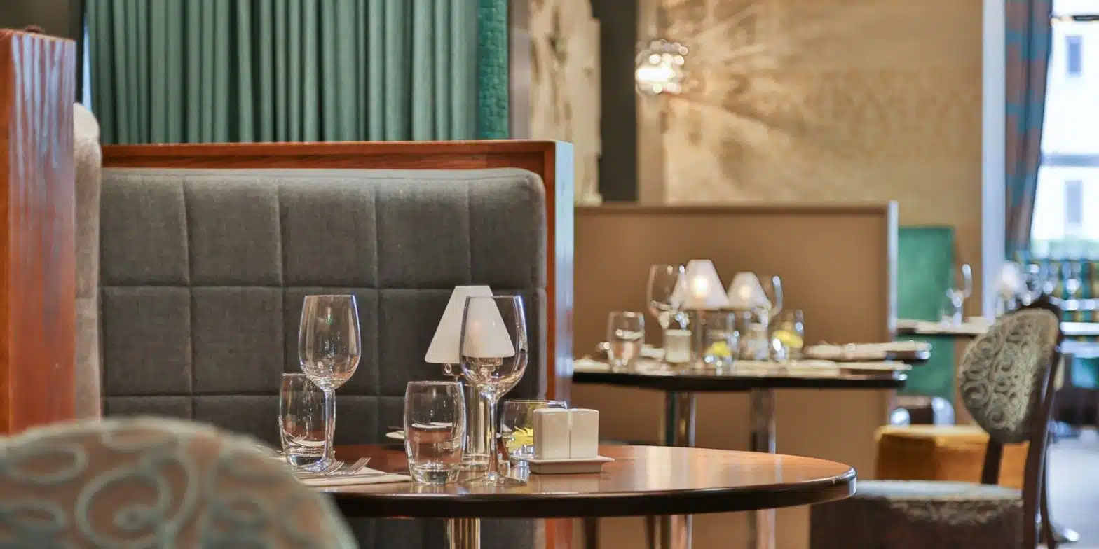 The westville hotel terrace restaurant dining area with set tables, featuring empty wine glasses, water glasses, small lamps, and upholstered chairs and banquettes. Westville Hotel Enniskillen menu