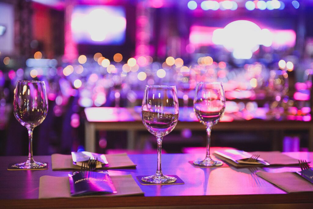 A close-up of wine glasses and table settings on a table, with blurred colourful lights and a crowd in the background.