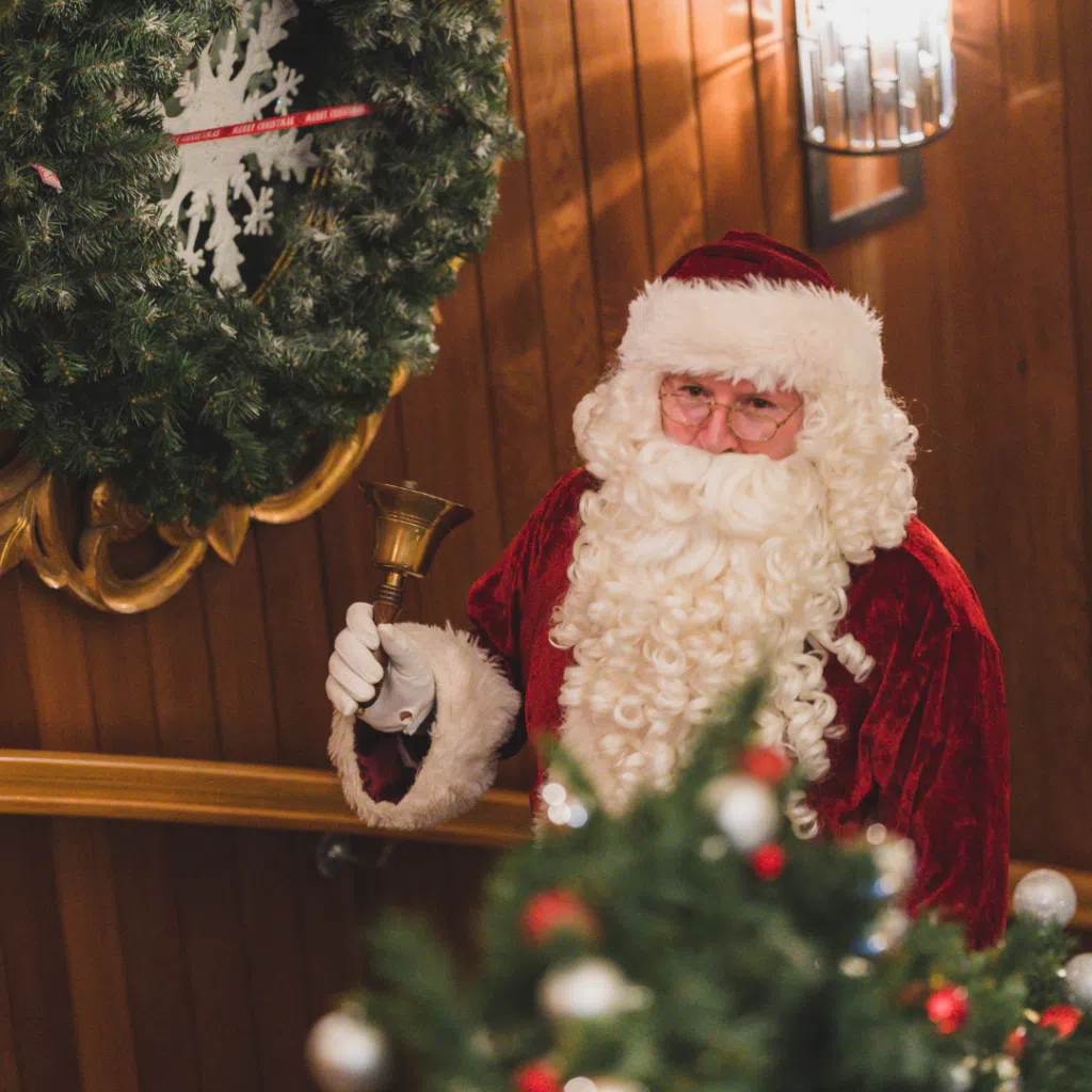 Santa Claus ringing a bell beside festive Christmas decorations and wreath indoors