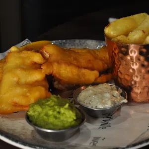 A plate of battered fish fillets with a side of chips in a copper cup, served with mushy peas and tartare sauce in small metal containers—a classic taste of Enniskillen dining. cosy autumn dining enniskillen