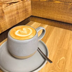 A grey ceramic cup of latte with heart-shaped latte art sits on a matching saucer with a spoon on a wooden table, offering a cosy touch to your Enniskillen Dining experience.