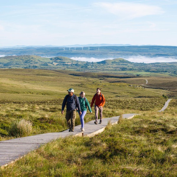 Three people walk on a wooden walkway through a grassy landscape with hills and a winding path in the background under a clear sky.