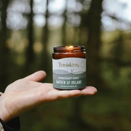 A hand holds a lit EnniskiNN aromatherapy candle labelled "Garden of Ireland" in a forest setting.