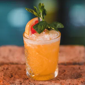 A glass of orange cocktail garnished with a grapefruit slice, mint leaves, and crushed ice sits on a textured surface—perfect for savouring during your Enniskillen dining experience.