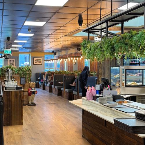 A modern café interior with wooden decor, booth seating along the wall, greenery accents, and a self-service counter in the foreground. Several people are seated and dining.