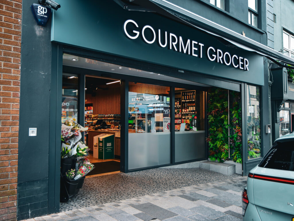 Street view of the entrance to a modern gourmet grocery shop with flowers displayed outside and shelves of products visible inside.