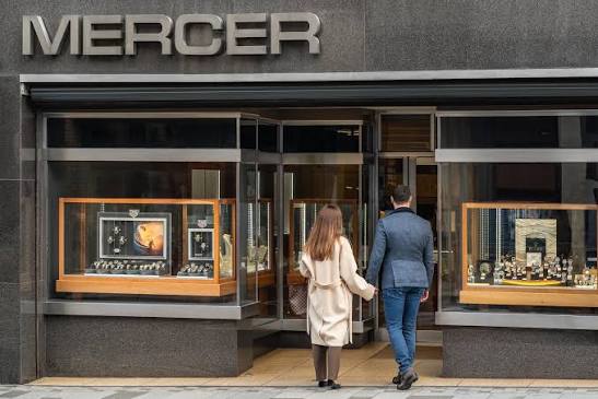 A couple holding hands walks into a shopfront labelled "MERCER" with watches and jewellery displayed in the windows.