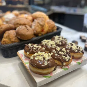A tray of chocolate-covered doughnuts topped with white and dark chocolate chips sits in front of a basket of scones on an Enniskillen Dining bakery counter.