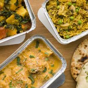 Three foil containers of Indian food, including curry, rice with vegetables, and palak paneer, served with naan bread on a wooden table— authentic Enniskillen Dining.