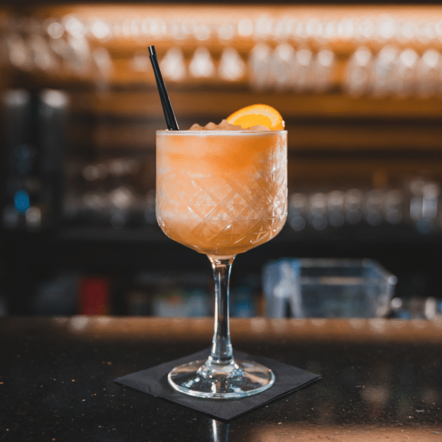 A cocktail with an orange slice garnish and a straw in a patterned glass sits on a bar counter with a black serviette, blurred glassware in the background.